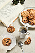 A relaxing setting with a book, sunglasses, a cup of black coffee, and freshly baked chocolate chip cookies on a vintage plate