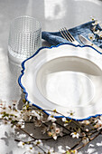 A sophisticated table setting featuring a white plate with blue detailing, glassware, and a sprig of spring blossoms on a textured surface