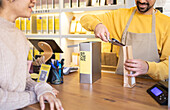 A cheerful barista in a yellow shirt scoops fresh coffee beans for a female customer at a cozy coffee shop.