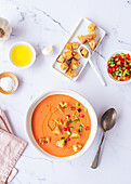 Top view of a vibrant bowl of traditional Spanish tomato gazpacho soup adorned with diced vegetables and golden croutons, served alongside ingredients on a marble surface.