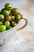 Close-up of fresh organic gooseberries in a rustic, tarnished metal cup set against a soft-focus background. The vibrant green and red tones of the berries highlight their ripeness and natural appeal.