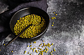 A rustic setting features raw organic mung beans in a textured black bowl, scattered loosely with a spoon, on a dark, grainy surface, highlighted by natural light