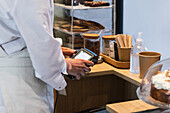 Side view of crop anonymous seller using POS terminal for payment while standing at counter in bakehouse with vegan desserts