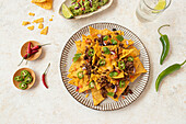 Top view of a savory plate of nachos piled high with beef, guacamole, melted cheese, jalapenos, and fresh toppings served alongside lime and refreshing drinks.