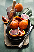 Juicy blood oranges halved and placed on a dark plate, with a green napkin and citrus juicer in the background