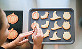 A person decorates freshly baked Halloween-themed cookies, including shapes like pumpkins and witch hats, on a grey baking tray