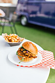 Eine Nahaufnahme eines köstlichen Gourmet-Burgers mit gewürzten Pommes frites, serviert auf einem karierten Papier, mit einem Food Truck im Hintergrund