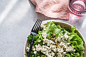 A nourishing herb salad mix featuring lettuce, parsley, and tarragon, topped with capers and crumbled blue brie cheese, accompanied by a glass of water