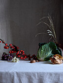 A tasteful arrangement featuring savoy cabbage, red grapes, and tiny red crab apples, artistically displayed on a draped white fabric with subtle lighting.