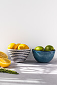 Two ceramic bowls, one with bright yellow lemons and the other with green limes, are set on a light table with soft shadows