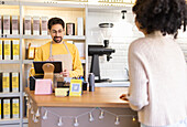 A cheerful business owner operates a cash register inside a cozy coffee shop while attending to a female customer.