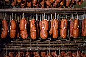 Reihen von Sobrasada, einer traditionellen Wurst von den Balearen, hängen in einem schwach beleuchteten Trockenraum und zeigen die satte rote Farbe und die strukturierte Außenseite, die mit weißen Schnüren gebunden ist.