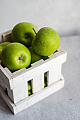 Vibrant green apples with a fresh dewy texture displayed in a rustic white wooden crate against a soft gray background