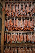 Rows of cured meats, prominently featuring traditional sobrasada, hanging in a rustic, dimly-lit drying room. These sausages are coated with a layer of white mold, indicative of the aging process.