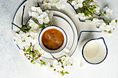 A top view of a cup of coffee surrounded by white spring blossoms and a milk jug, set on a white backdrop