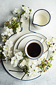 A cup of black coffee and fresh milk in a white enamel mug alongside cherry blossoms on a plate, capturing the essence of spring