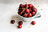 Bowl filled with juicy red cherries, some scattered on a textured white background.