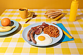 In a coloured Tablecloth lays a blue plate containing a delicious Breakfast with Eggs, beans and sausages, and there are some bread slices, a cup of coffee and some fruit