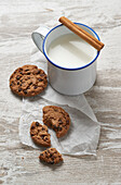 A rustic mug of milk garnished with a cinnamon stick beside freshly baked chocolate chip cookies on a weathered wooden table