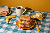 Ein hausgemachtes Frühstück auf einer karierten blauen Tischdecke mit einem Sandwich, einer Banane, Kaffee und Butter vor gelbem Hintergrund