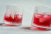 Two textured glasses filled with ice and a vibrant cherry vodka and tonic cocktail, cast on a simple backdrop.