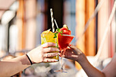 Two cropped unrecognizable friends clinking glasses, one holding a vibrant mango mojito, the other a frosty strawberry cocktail, symbolizing celebration and joy.