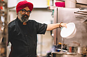 An experienced chef wearing a red turban pours ingredients into a steaming pot in a professional kitchen setting