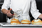 Unrecognizable crop chef caramelizing sugar cream on sweet Berliners with gas torch while preparing vegan dessert in bakehouse