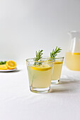 Two glasses of freshly made lemonade with rosemary sprigs and a sliced lemon in the background on a clean white surface