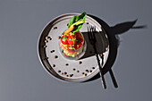 Top view of a colorful vegetable salad neatly presented in a transparent glass, placed on a textured gray plate. The arrangement includes fresh greens, cherry tomatoes, and a carrot, accompanied by a fork and scattered chickpeas around the plate.