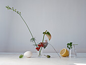 A serene arrangement featuring a twig, egg, lemon, currant and a sprig in glass vases on a white surface, embodying simplicity and natural beauty