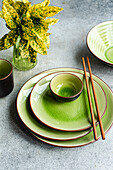 From above view of a modern table setting featuring bright green ceramic dinnerware and wooden chopsticks, juxtaposed with a vibrant potted plant, all showcased on a textured grey background.