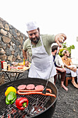 A man in a chef's hat and apron grills food at an outdoor BBQ party, looking down as he cooks. Two friends sit nearby, engaging in conversation and enjoying the atmosphere.