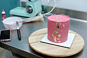 A sophisticated pink cake adorned with edible gold leaf, showcased on a wooden board, with baking tools in the background