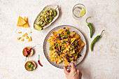 Cropped unrecognizable person's hand reaching for tasty nachos topped with cheese, guacamole, and jalapenos, captured in a bright setting with refreshing drinks on the side.