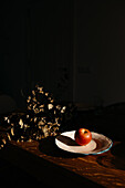 A solitary apple rests on a plate illuminated by a sunbeam against a dark backdrop, with a bouquet of dried flowers adding texture to the scene