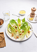 Top view of a Caesar salad with crisp lettuce, golden croutons, and shaved Parmesan on a white plate. Salt, pepper, a wooden grinder, crouton bowl, lemon wedge, and dressing bowl are also visible.