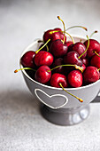 From above juicy red cherries in a charming ceramic bowl with heart motifs, set against a soft gray backdrop. The vibrant red of the cherries contrasts beautifully with the muted tones of the bowl and background, emphasizing freshness and flavor.