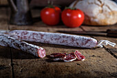 A rustic still life featuring fuet and another type of cured sausage, fresh tomatoes, and crusty bread on an old wooden table, emanating a traditional culinary charm.