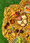 Eine einladende Sommerpicknick-Szene mit einer Vielzahl von Snacks und Früchten, die wunderschön auf einer bunten, blumengemusterten Decke auf üppigem grünen Gras arrangiert sind. Dazu gehören Käse, Fleisch, Weintrauben und Pfirsiche