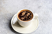 A From above of a freshly brewed cup of black coffee, with visible bubbles, served in a white ceramic cup seated on a matching saucer, set on a textured grey surface.