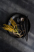 Top view of sophisticated black dining setup featuring a plate, utensils, and a pop of color from bright yellow mimosa flowers against a dark textured background.