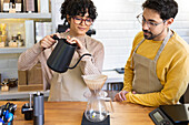 Zwei multiethnische Baristas arbeiten zusammen, um Pour-over-Kaffee aufzubrühen, wobei sie Teamarbeit und Geschicklichkeit in einer warmen, einladenden Caféumgebung demonstrieren.