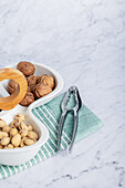 From above white ceramic bowl filled with whole walnuts beside a bowl of pistachios, with a walnut cracker and a decorative wooden band. Set on a marble surface with a striped green and white towel, this setup exemplifies wholesome snacking options.