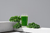 Vibrant green kale juice in a transparent glass, accompanied by fresh kale leaves, elegantly placed on a white platform with a neutral background