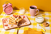 Colorful breakfast scene featuring waffles topped with strawberries and whipped cream, served on a pastel pink plate with coffee and milk on a yellow checkered tablecloth