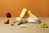 Top view of assorted cheeses alongside fruits and nuts on a neutral-toned surface. The composition includes brie, cheddar, and swiss, complemented by grapes, a plum, and a peach