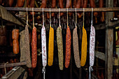 A selection of cured sausages, including sobrasada, hangs in a rustic smokehouse. The sausages are seasoned with various spices, showcasing a range of colors from deep red to yellow.