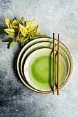 From above, a neatly arranged table setting featuring vibrant green ceramic plates and brown chopsticks, accompanied by a decorative plant on a textured surface.