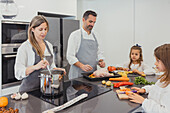 Eine Familie arbeitet harmonisch in einer modernen Küche und bereitet ein gebratenes Hühnchen mit einer Reihe von buntem Gemüse zu, was eine herzerwärmende häusliche Szene zeigt